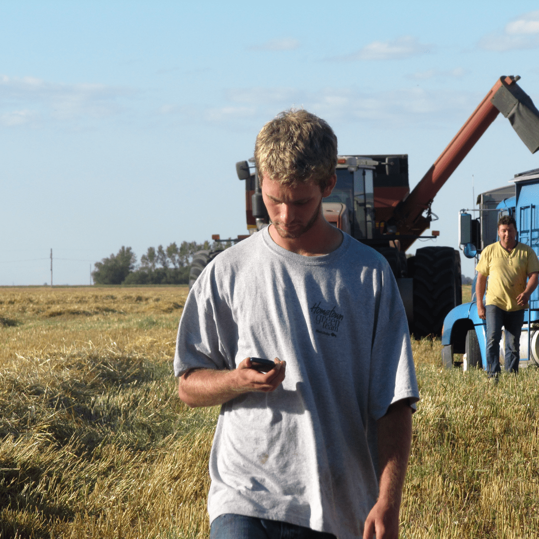 Home Page Image - AgChieve A young farmer looks at his phone walking away from a truck driver in the field.